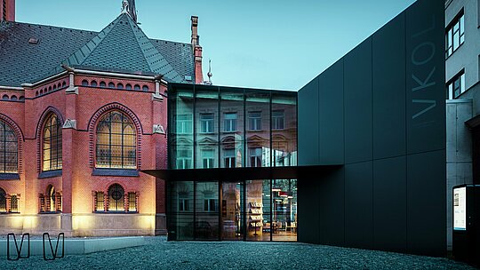 Obrázok ukazuje pôsobivé spojenie historickej a modernej architektúry v Olomouci. Výrazný Červený kostol v gotickom štýle s umeleckými oknami a vysokou vežou dopĺňa moderná novostavba s minimalistickou čiernou fasádou z hliníkových kompozitných dosiek PREFABOND. Transparentné sklenené plochy novostavby odrážajú historické prostredie a vytvárajú fascinujúci kontrast s masívnou štruktúrou kostola. Presné spracovanie moderných materiálov v kombinácii s harmonickým spojením starého a nového dodáva budove modernú eleganciu a zároveň rešpektuje históriu okolia. Táto kombinácia inovatívnych a odolných materiálov, ako je PREFABOND, zaručuje estetickú krásu aj funkčnosť.