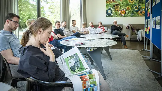 PREFARENZEN 2027 Diskusia odborníkov v svetlej seminárnej miestnosti: Paulina Frey (vedúca marketingu PREFA CH) drží v rukách veľkú knihu o architektúre s projektmi z hliníka PREFA, zatiaľ čo ostatní členovia poroty pozorne počúvajú. Veľké okná vpúšťajú dovnútra denné svetlo a ponúkajú výhľad do zelene. Scéna vyžaruje sústredenú analýzu, kreatívnu diskusiu a hodnotenie dizajnu a funkčnosti. Hliníkové produkty PREFA symbolizujú trvácne, estetické strechy a fasády, ktoré podporujú inováciu, kvalitu a inšpiráciu v architektonických projektoch.