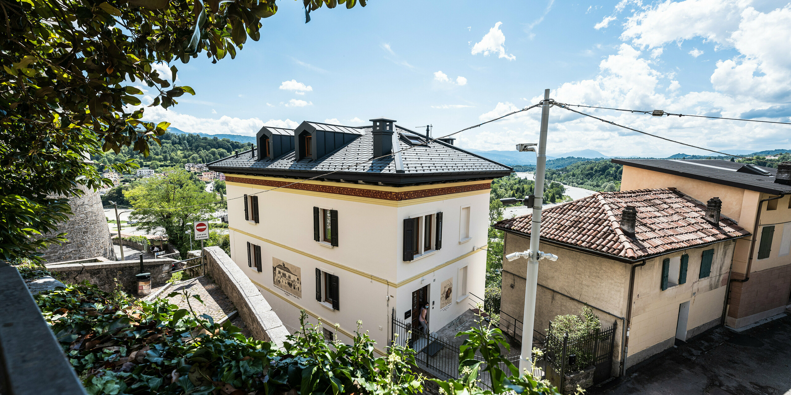 Panoramatický pohľad na zrekonštruovanú historickú obytnú budovu v talianskom meste Belluno, zasadenú do mestského prostredia s výhľadom na zelené kopce. Elegantnú fasádu v klasickom štýle zo začiatku 20. storočia dopĺňa hliníková strecha PREFA so šindľom antracitovej farby, ktorá predstavuje moderné a udržateľné riešenie. Jemné začlenenie vikierov a komínov PREFALZ vo farbe P.10 antracit poskytuje príťažlivý vzhľad a odráža snahu o zachovanie historickej architektúry.