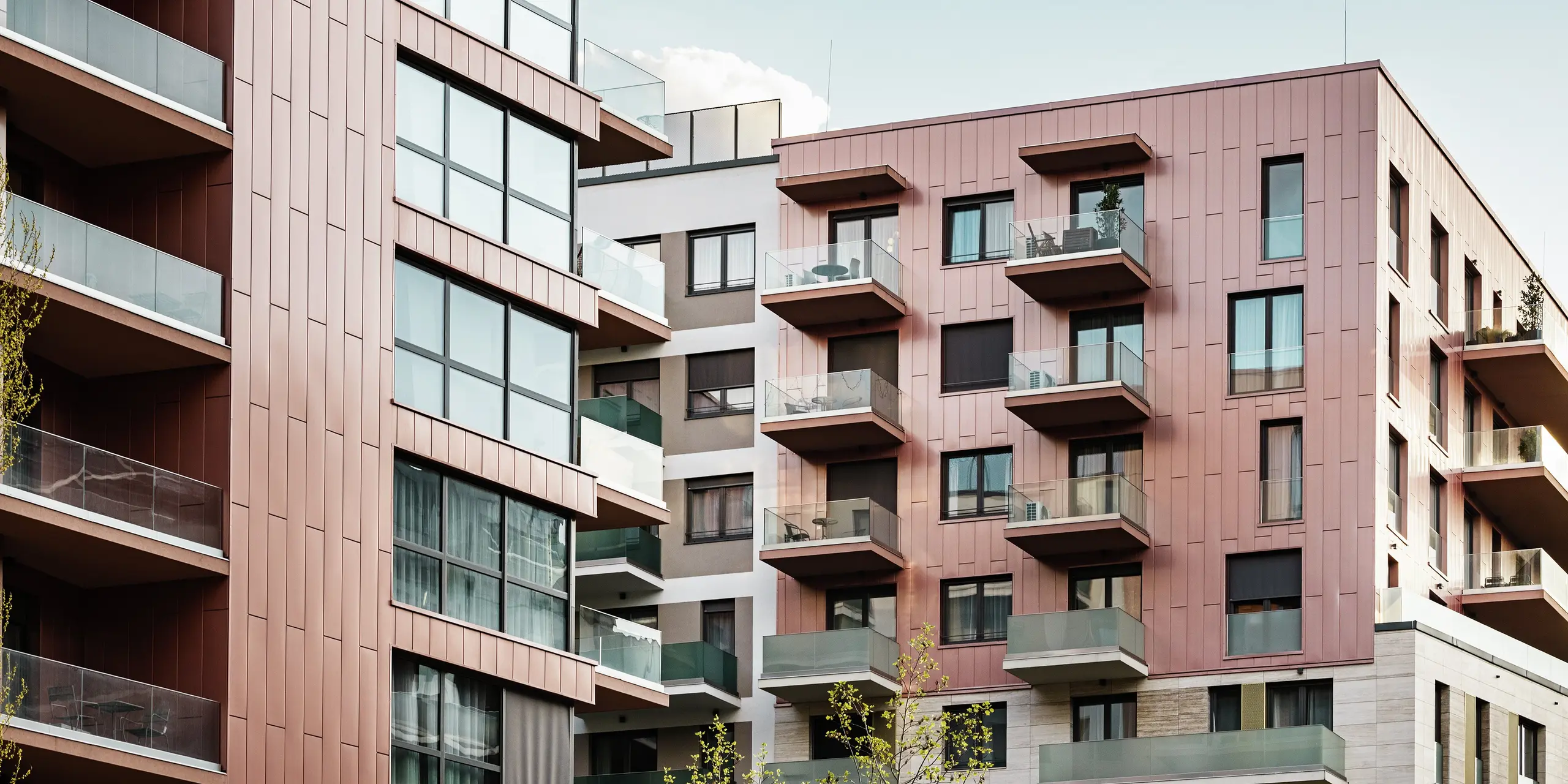 Detail fasády obytného komplexu Grand Corvin v revitalizovanej štvrti Corvin v Budapešti. Vertikálne položené fasádne panely PREFA Siding v špeciálnej farbe lososová metalíza dotvárajú moderný vzhľad horných poschodí budovy. V kombinácii s veľkoplošnými sklenenými fasádami a prírodným kameňom vytvára odolný hliníkový obklad harmonický a kvalitný obraz mesta.
