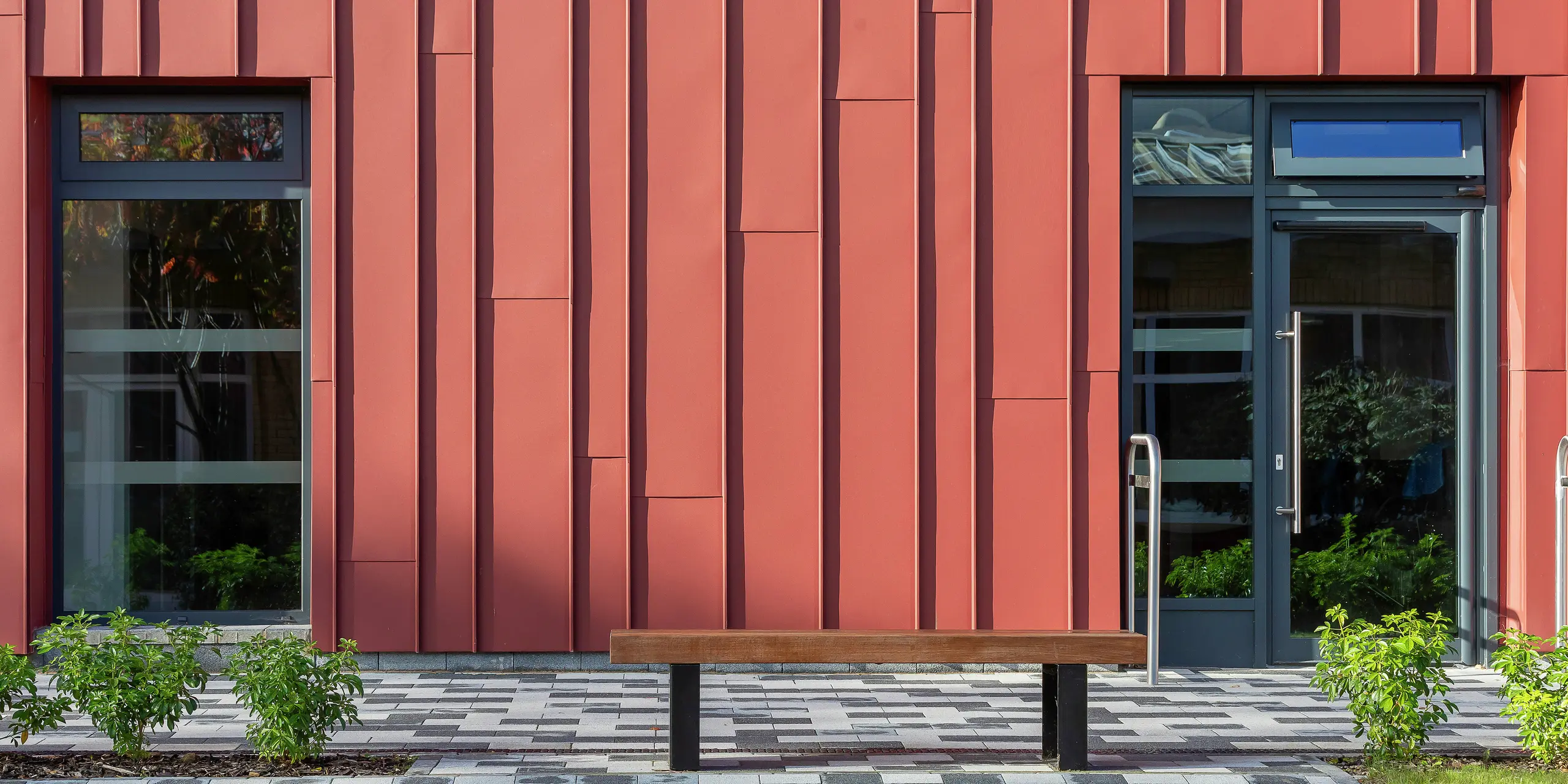 Detailný dizajn fasády s PREFALZ: Obklad so stojatým falcom s PREFALZ v odtieni P.10 tmavočervená dodáva Soldiers' Centre v Pirbright moderný a dynamický vzhľad. Okná a dverové rámy v P.10 Anthrazit vytvárajú elegantné kontrasty, presná montáž zaisťuje jedinečnú hru fasády. Vysoko kvalitné hliníkové produkty PREFA pre trvácnu a estetickú architektúru.