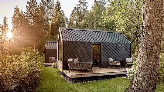 Tiny House v areáli Secret Koi Garden je zasadený do pokojnej lesnej krajiny pri jazere Natterer See a je príjemne osvetlený nízko stojacim večerným slnkom. Strecha a fasáda sú obložené malými hliníkovými šablónami PREFA v odtieni P.10 antracitová, ktorých štruktúrovaný povrch pripomína rybí kožúšok. Robustný a odolný hliníkový plášť spoľahlivo chráni pred poveternostnými vplyvmi a podčiarkuje čistú, nadčasovú architektúru glampingu uprostred prírody.