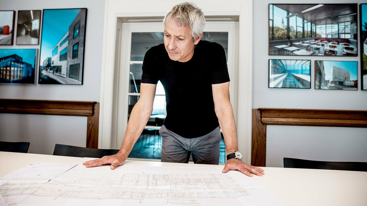 Architekt pracuje na podrobných stavebných plánoch v modernej kancelárii, obklopený pôsobivými fotografiami architektonických projektov PREFA.