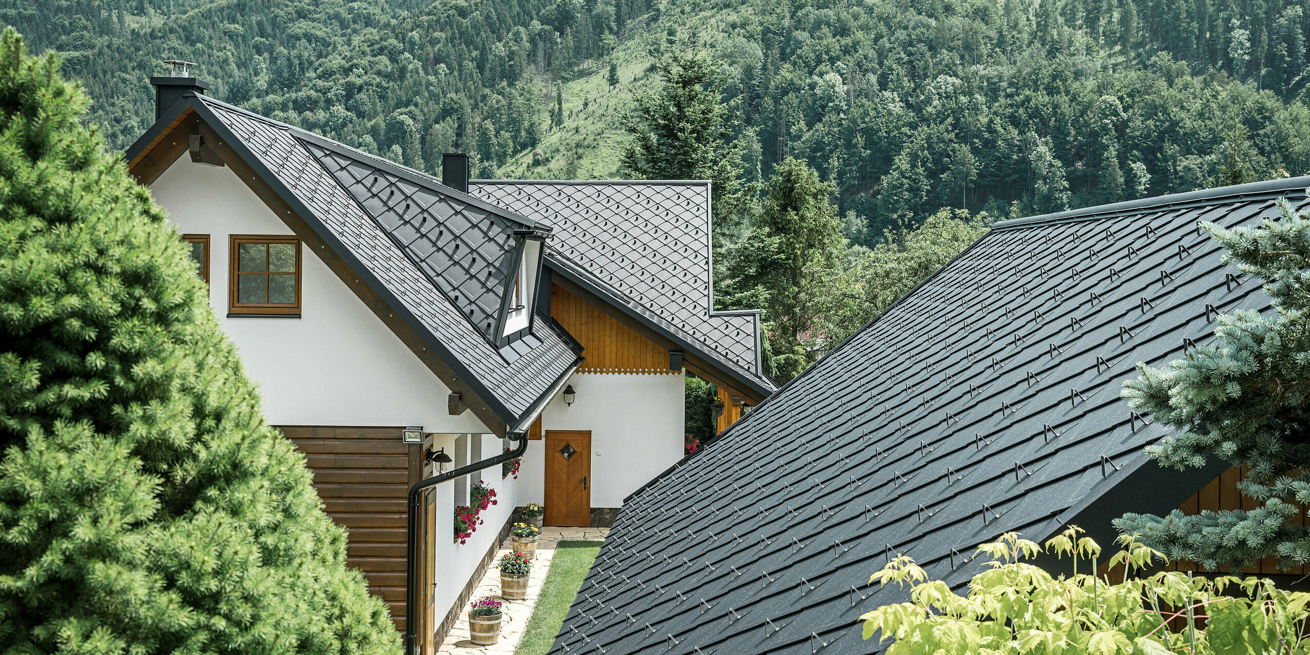 Zadný pohľad na tradičný rodinný dom v Starej Bystrici na Slovensku s úchvatnou strešnou krajinou zo šablón PREFA 29 × 29 v odtieni P.10 čierna. Robustné hliníkové šablóny dokonale harmonizujú s prírodným prostredím a ponúkajú moderný kontrast k zalesnenej kopcovitej krajine v pozadí. Detailne prepracované strešné plochy a dobre zakomponované trapézové vikiere dodávajú budove jedinečný estetický vzhľad.