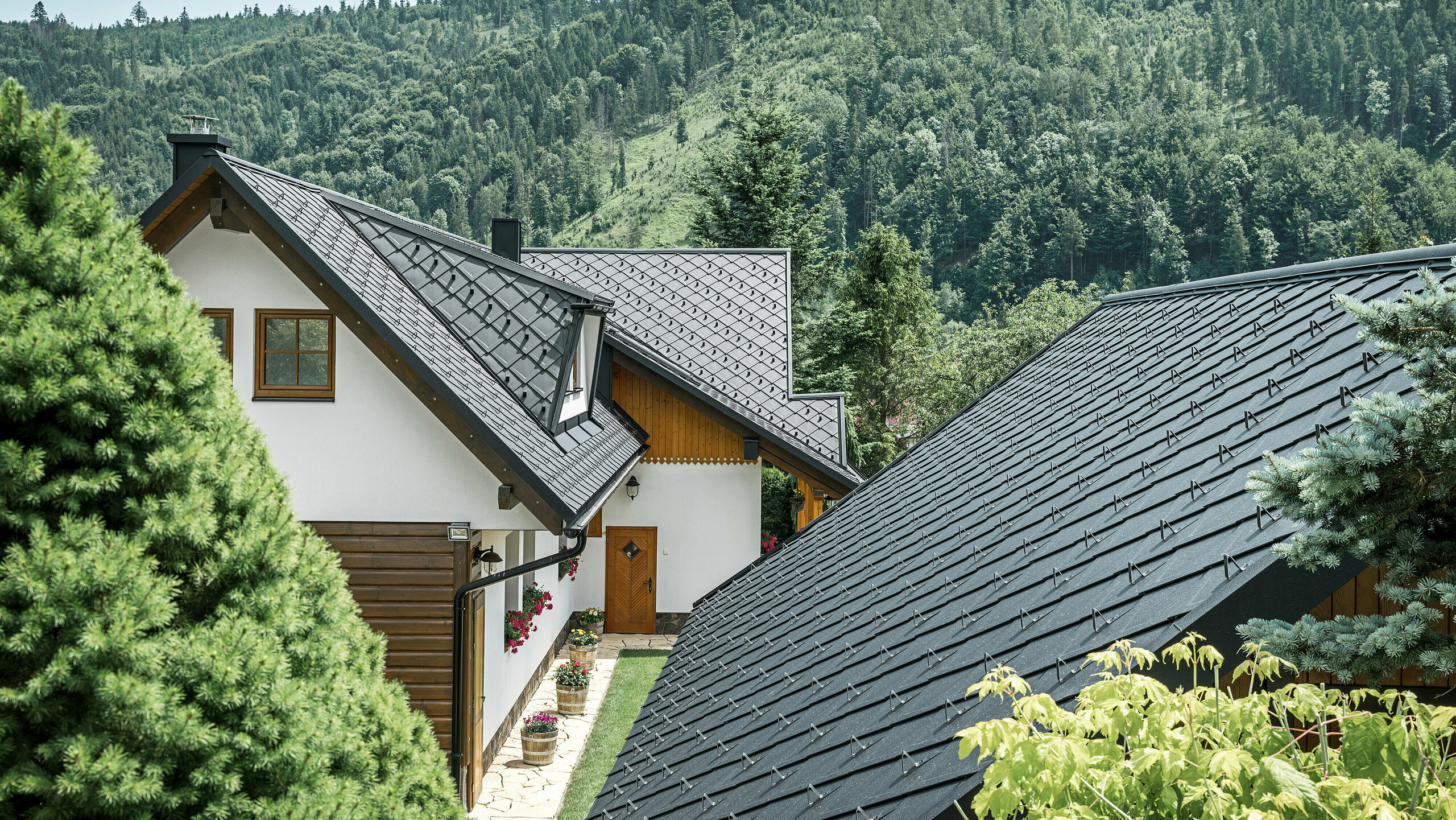 Zadný pohľad na tradičný rodinný dom v Starej Bystrici na Slovensku s úchvatnou strešnou krajinou zo šablón PREFA 29 × 29 v odtieni P.10 čierna. Robustné hliníkové šablóny dokonale harmonizujú s prírodným prostredím a ponúkajú moderný kontrast k zalesnenej kopcovitej krajine v pozadí. Detailne prepracované strešné plochy a dobre zakomponované trapézové vikiere dodávajú budove jedinečný estetický vzhľad.