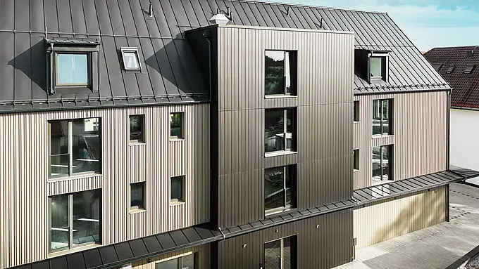 Na snímke je moderná budova vo Feldkirchene, ktorú navrhol architekt Stefan Heigl. Fasáda je obložená materiálom Prefalz vo farbe bronzová, zatiaľ čo šikmá strecha je pokrytá materiálom Prefalz v čiernošedej farbe. Veľké okná a strešné okná poskytujú v budove dostatok prirodzeného svetla. V návrhu sa spájajú vysokokvalitné materiály a elegantná architektúra, ktorá je funkčná aj estetická. Tento moderný spôsob výstavby a použitie materiálov Prefalz dodávajú budove moderný a štýlový vzhľad.