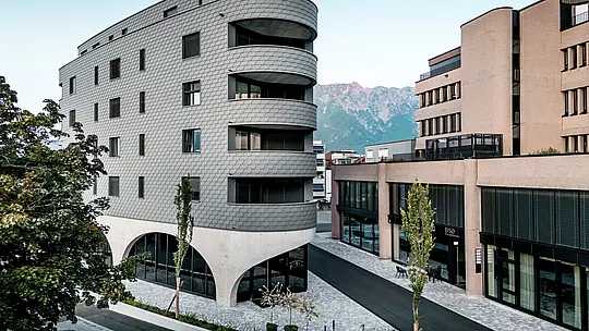 Pohľad na Bernerhaus v Buchsi s výrazným trojuholníkovým tvarom a jemne zaoblenými balkónmi pred alpskou horskou kulisou. Súvislá fasáda z PREFA fasádnych šindľov v odtieni P.10 svetlošedá obklopuje budovu ako šupinatá hliníková koža a vytvára živú hru svetla a tieňa. Nad masívnym betónovým podstavcom s elegantnými arkádami pôsobí ľahká, odolná proti poveternostným vplyvom PREFA hliníková fasáda mimoriadne precízne, trvalo a s nízkymi nárokmi na údržbu – architektonický pohľad v mestskom prostredí.