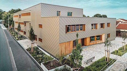 Večerná atmosféra ponára detské jasle v Alzenau do jemného svetla a zvýrazňuje kvalitnú fasádu z bronzovo sfarbených PREFA fasádnych šablón 44 × 44. Kombinácia štruktúrovanej hliníkovej fasády a vertikálnych drevených plôch vytvára živú hru materiálov, farieb a textúr. Veľkorysé okná zabezpečujú svetlé interiéry, zatiaľ čo okolité zelené plochy a vydláždené predpolie pozývajú k pobytu. Obrázok pôsobivo ukazuje, ako súčasná architektúra, funkčnosť a udržateľnosť harmonicky realizované s produktmi PREFA – priamo v mestskom kontexte Alzenau.