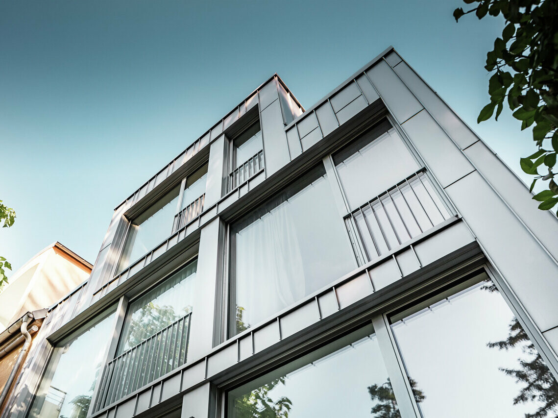 Pohľad zospodu na moderný bytový dom s jasnou geometrickou architektúrou, charakteristickou lesklou striebornou metalickou hliníkovou fasádou Prefalz. Veľké priečelia okien odrážajú modrú oblohu a okolitú vegetáciu. Budovu charakterizujú úzke balkóny s kovovým zábradlím, ktoré podčiarkujú minimalistický dizajn.