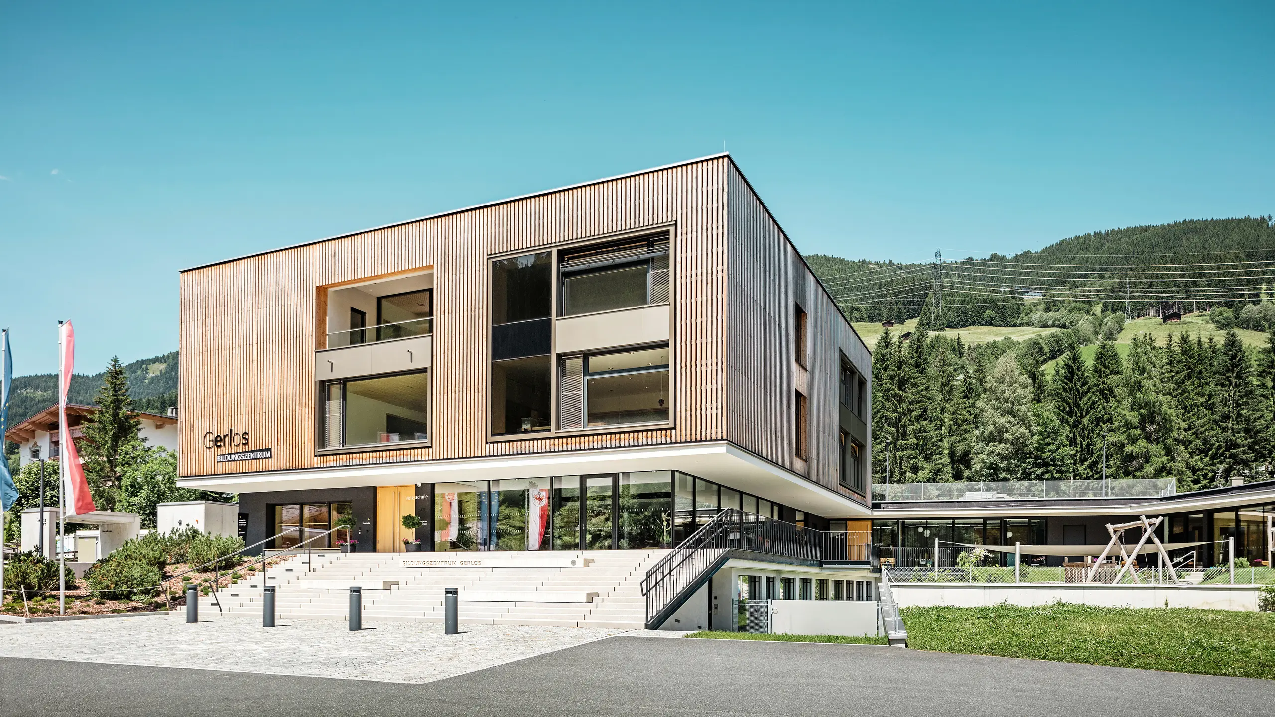 Pohľad na moderné vzdelávacie centrum s drevenou fasádou spredu. Budova je dvojpodlažná s veľkými okennými priečeliami a plochou strechou, ktorá dokonale zapadá do okolitej horskej krajiny husto zalesnených kopcov. V popredí sú upravené zelené plochy, stupňovitá príjazdová cesta a reklamné vlajky. Návrh vyjadruje kombináciu funkčnosti a estetickej architektúry v harmonickom súlade s prírodou.