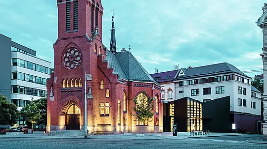 Obrázok ukazuje harmonické spojenie historickej a modernej architektúry v Olomouci. Impozantný Červený kostol s výraznou špicatou vežou a gotickými oknami tvorí historické centrum komplexu. Priamo vedľa neho sa týči moderná novostavba, ktorej fasáda z matných hliníkových kompozitných dosiek PREFABOND a skla vytvára jasný, elegantný kontrast s tradičnou architektúrou. Veľkoplošné sklenené fasády poskytujú pohľad do novej budovy, ktorá je inovatívnym spôsobom prepojená s kostolom. Moderná budova svojimi presnými tvarmi a čistými líniami prispieva k súčasnej atmosfére, bez toho aby narušila historický charakter kostola. Táto vydarená symbióza ukazuje, ako vysoko kvalitné materiály, ako je PREFABOND, poskytujú perfektný základ pre spojenie tradície a modernosti.