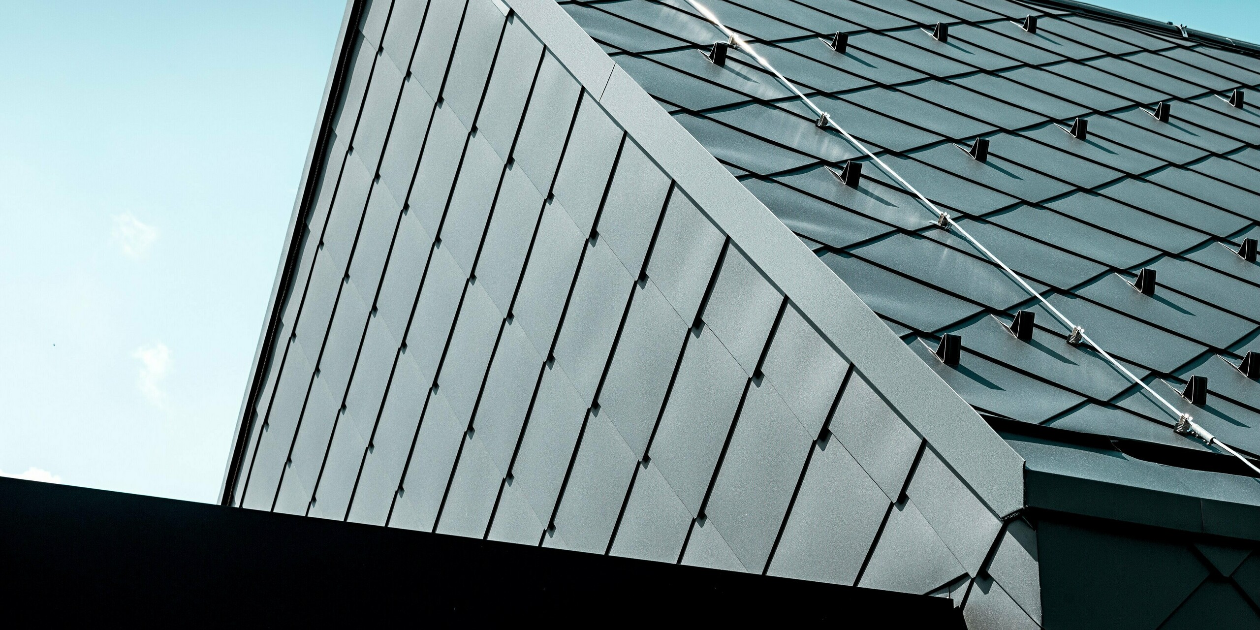 Na obrázku je zachytený výrazný detail rodinného domu v Trnave, ktorého strecha je kompletne pokrytá strešnými šablónami PREFA 44 × 44 v odtieni P.10 tmavošedá. Presne položená štruktúra šablón a integrované snehové zábrany zdôrazňujú vysokú kvalitu spracovania a technickú funkčnosť hliníkovej krytiny. Matný povrch rovnomerne odráža svetlo a podčiarkuje čistý, moderný tvar štítu. Odolné produkty PREFA ponúkajú optimálnu ochranu, vysokú stabilitu a nadčasový elegantný vzhľad