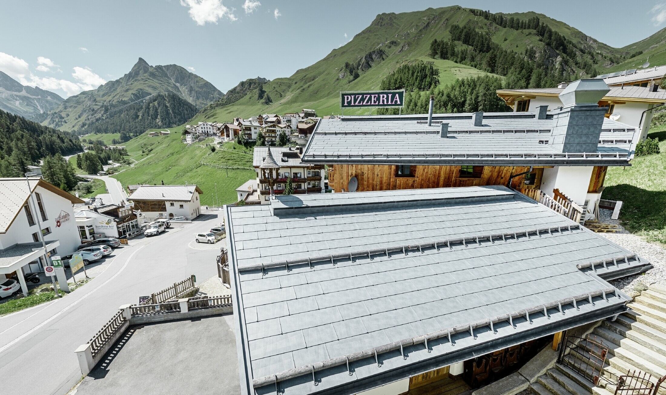 Reštaurácia Almrausch s horami v pozadí so strechou PREFA zo strešných panelov FX.12 vo farbe kamenná šedá s horskými protisnehovými zábranami