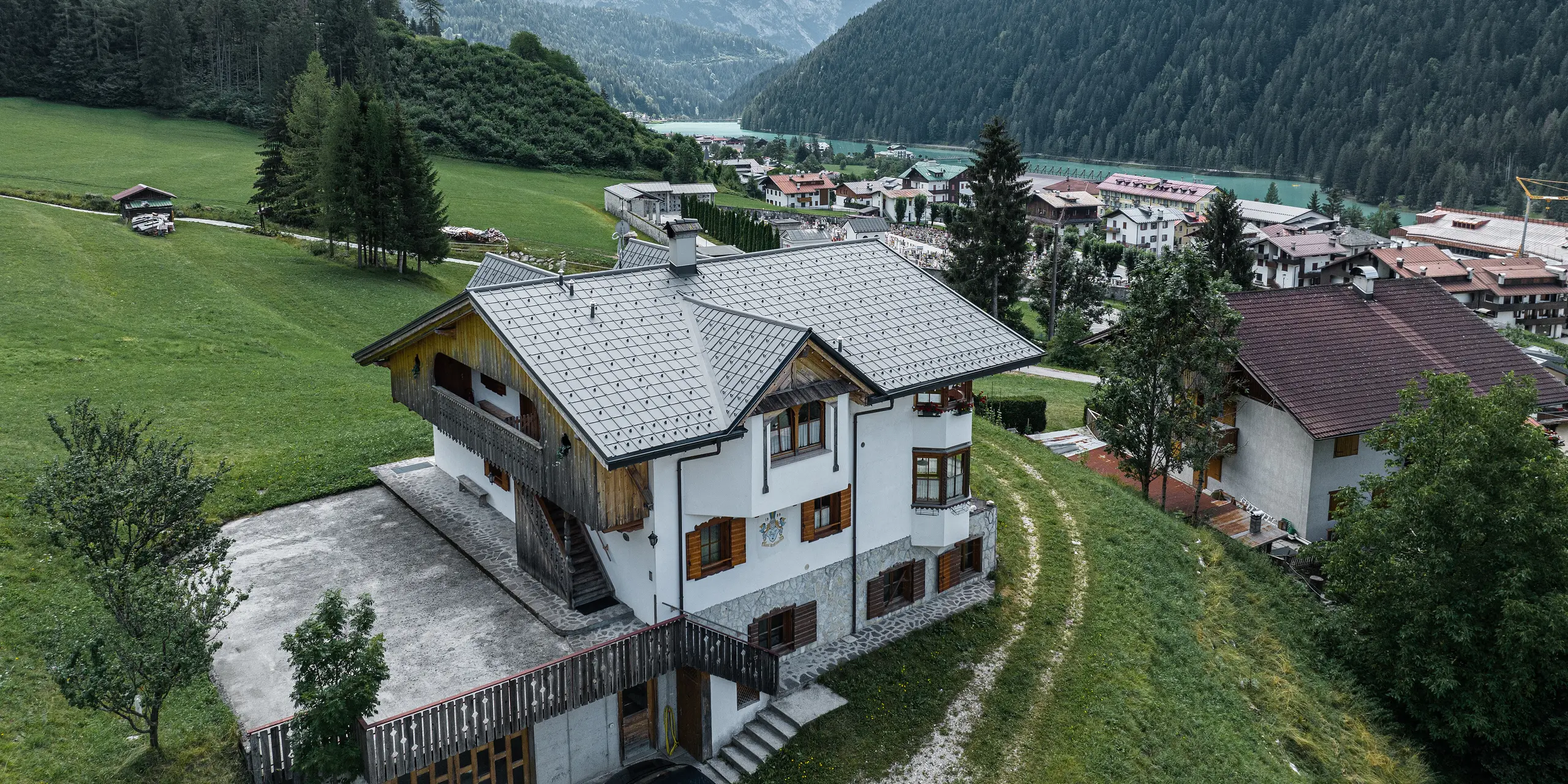 Letecký pohľad na pôvabnú horskú chatu v Auronzo di Cadore v Taliansku po renovácii strechy s hliníkovými škridlami PREFA v svetlosivej farbe. Chata sa týči na zelenej lúke s pôsobivým horským masívom v pozadí a ukazuje malebnú súhru modernizovaných stavebných prvkov a tradičnej alpskej architektúry. Nad vstupným priestorom je vidieť špicatý vikier s dreveným štítom.