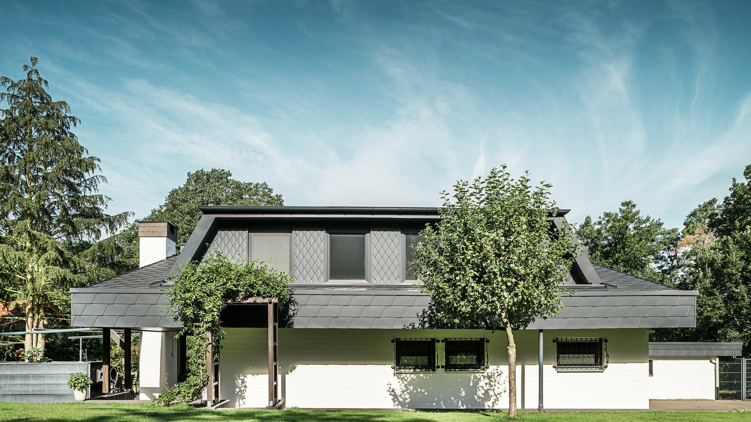 Pohľad spredu na záhradnú stranu rodinného domu v Mechernichu s veľkým trapézovým vikierom, pokrytým malými antracitovými strešnými šablónami PREFA P.10. Strecha je pokrytá šindľom PREFA DS.19 v rovnakej farbe. Fasáda spodného podlažia je natretá na bielo a celkový obraz v záhradnej časti dotvára zelená pergola. Stromy a kríky rámujú výhľad a zabezpečujú harmonické začlenenie do prírody.