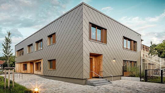 Moderná materská škola v Alzenau zaujme svojou čistou architektúrou a kvalitnou hliníkovou fasádou PREFA v bronzovej farbe. Fasáda z hliníkových šablón 44 × 44 dodáva budove elegantný, štruktúrovaný vzhľad a vizuálne spája jednotlivé funkčné časti do harmonickej jednoty. Drevené obklady pri vstupoch vytvárajú teplý, prirodzený kontrast k trvácnemu hliníku. Kombinácia masívnej stavby, drevených prvkov a zavesenej prevetrávanej fasády je príkladom udržateľného stavebného riešenia. Nenáročný hliníkový plášť PREFA presviedča nielen svojou recyklovateľnosťou, ale aj odolnosťou voči poveternostným vplyvom – ideálny pre perspektívne vzdelávacie stavby s vysokými architektonickými nárokmi.