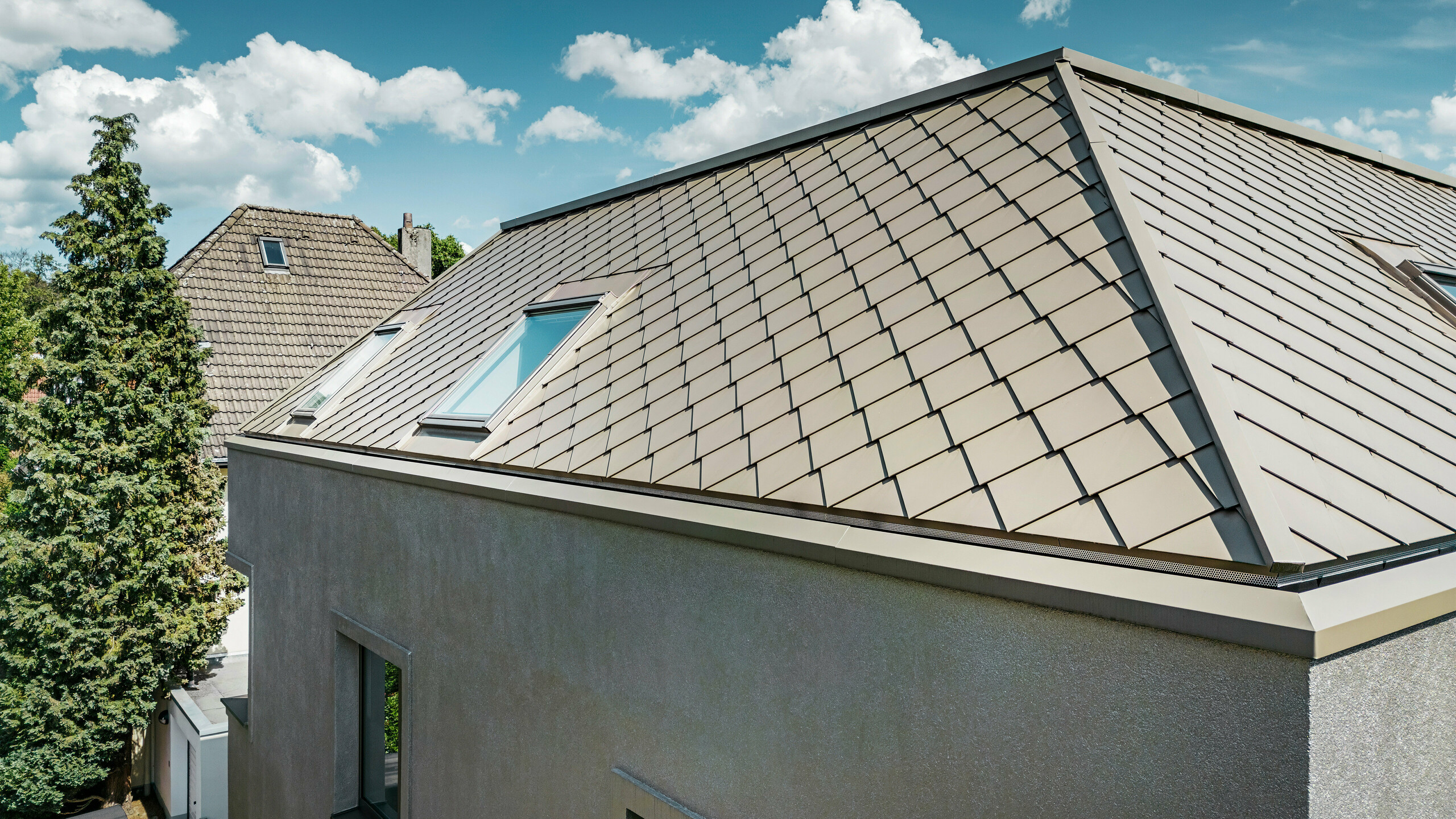 Detailný záber na valbovú strechu moderného rodinného domu s hliníkovým strešným plášťom bronzovej farby od spoločnosti PREFA. Strešná konštrukcia vykazuje vysokokvalitnú povrchovú úpravu s integrovanými strešnými oknami, ktoré poskytujú dodatočné prirodzené svetlo v interiéri. Čisté línie a lesklý povrch hliníkových strešných panelov zdôrazňujú moderný a elegantný dizajn domu.