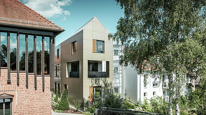 Na obrázku je moderná radnica v Alzenau, ktorej fasáda je vyrobená z hliníkových kompozitných dosiek PREFA v bronzovej farbe. Špicatá strecha a jasne štruktúrované okná s drevenými žalúziami dodávajú budove elegantný a moderný vzhľad. V popredí je vidieť zelená plocha s dláždenou cestičkou a stromami, ktorá harmonicky dopĺňa mestský dizajn budovy.
