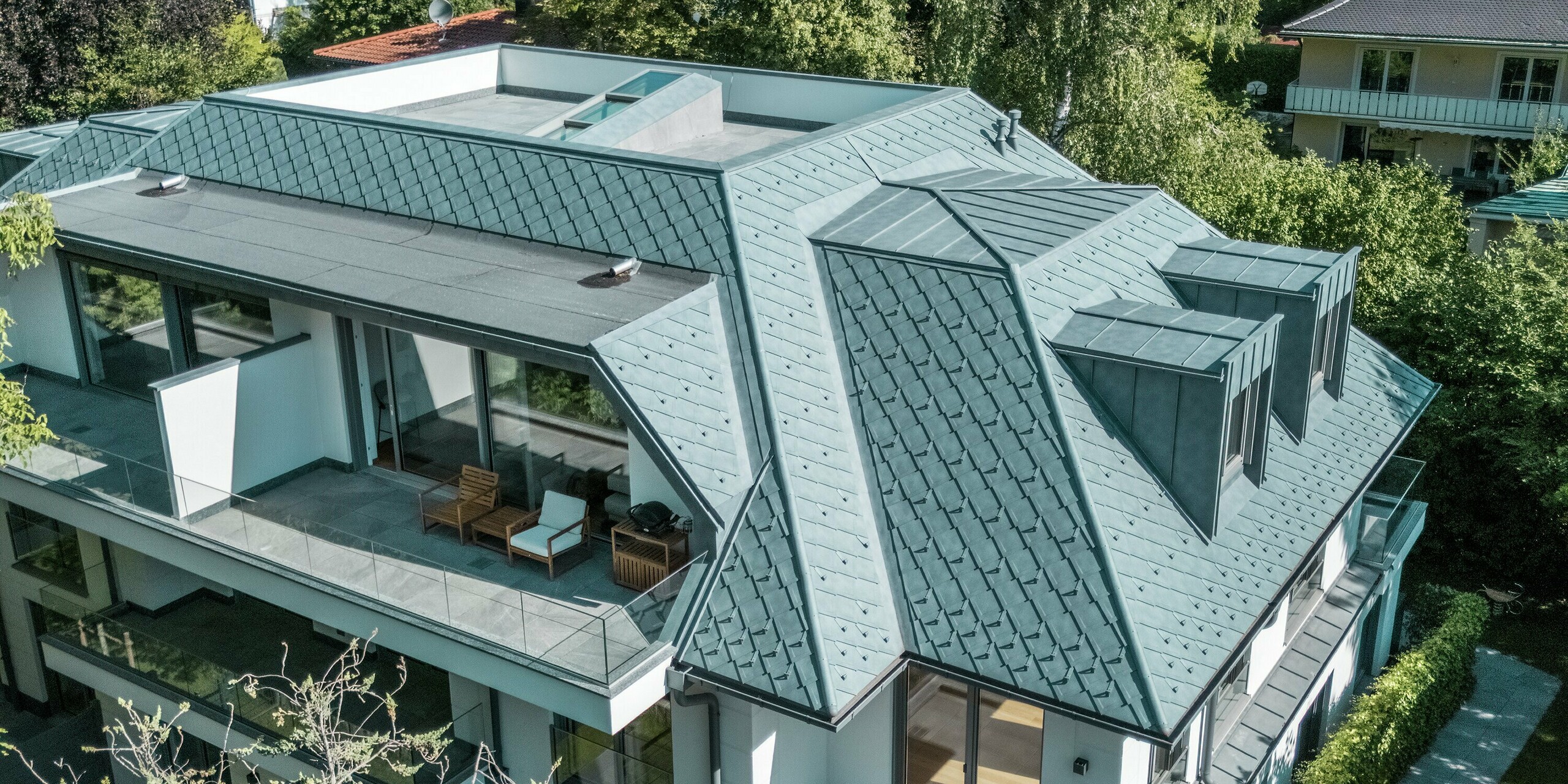 Luftaufnahme eines modernen Mehrfamilienhauses, dessen Dach mit PREFA Dachrauten 29 × 29 in der eleganten Farbe P.10 Steingrau gedeckt ist. Das prägnante, strukturierte Dachdesign wird durch die stilvollen PREFALZ Flachdachgauben ergänzt, die harmonisch in die Architektur integriert sind. Die großzügigen Terrassenflächen und die grüne Umgebung unterstreichen das elegante Gesamtbild. Die Verwendung hochwertiger Aluminiumprodukte sorgt für eine langlebige und witterungsbeständige Lösung, die sowohl ästhetisch als auch funktional überzeugt.