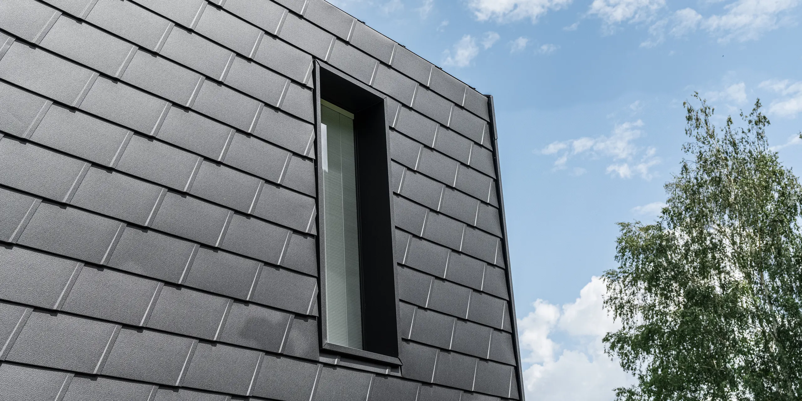 Detailný pohľad na fasádu s fasádnymi šindľami PREFA v čiernej farbe, ktoré rámujú štíhle vertikálne okno. Šindle poskytujú štruktúrovaný, robustný povrch, ktorý esteticky kontrastuje s prírodným prostredím a modrou oblohou.