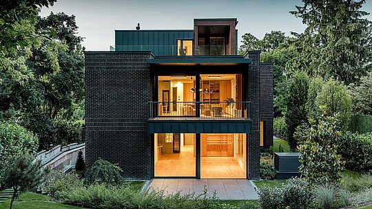 Fotografia zachytáva modernú zadnú stranu budapeštianskej viacgeneračnej vily za súmraku. Veľkorysé presklené fasády ponúkajú výhľad na príjemne osvetlené interiéry, zatiaľ čo kombinácia tmavočervenej tehlovej fasády a hliníka PREFALZ v odtieni P.10 antracit zdôrazňuje čisté architektonické línie. Presne vyhotovené uhlové stojaté falce podčiarkujú vysokú kvalitu spracovania a dlhú životnosť hliníkových produktov PREFA. Obklopená bujnou zeleňou sa budova harmonicky začleňuje do záhradnej krajiny a vyžaruje elegantný, moderný charakter.
