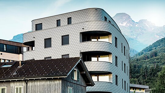 Moderná trojuholníková obytná a obchodná budova v Buchsi s výraznou zaoblenou architektúrou a šupinatou fasádou z PREFA fasádnych šindľov v odtieni P.10 svetlošedá. Rovnomerná hliníková fasáda vytvára živý dojem plazov a ako elegantný pohľadový prvok vyniká na pozadí alpských vrcholov. Vďaka ľahkému, odolnému voči poveternostným vplyvom a trvácnemu hliníku PREFA sú presne realizované aj úzke polomery a zaoblenia – pre homogénny, nenáročný na údržbu plášť budovy s nadčasovým moderným vzhľadom.