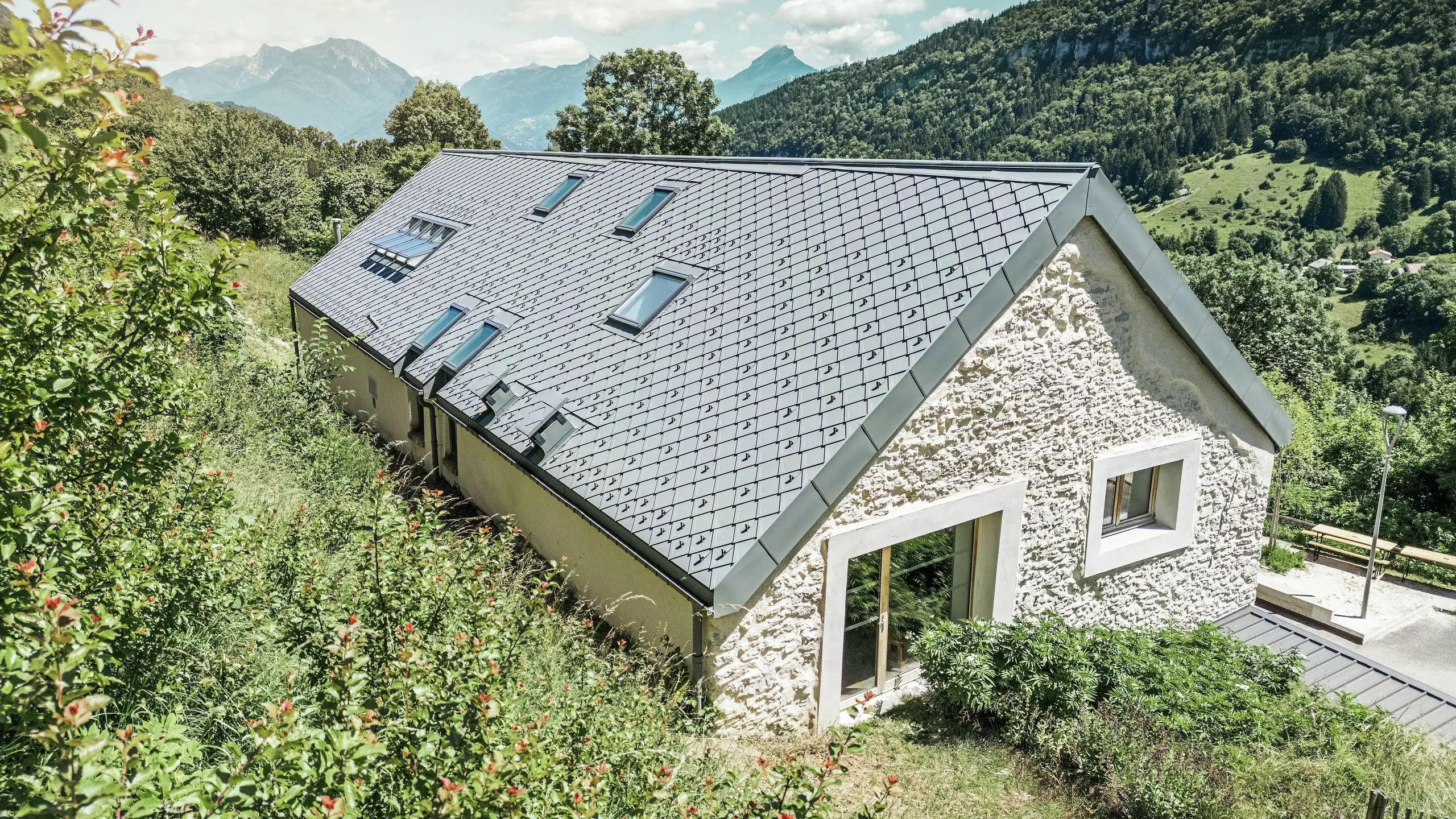 Pohľad na strechu zrekonštruovanej stodoly „La Grange à Gaby“ v Engins, Francúzsko. Budova je pokrytá odolnými strešnými šablónami PREFA 29 × 29, ktoré spájajú modernú ochranu s tradičnou estetikou. Idylická horská krajina zdôrazňuje harmonické začlenenie do prírody.