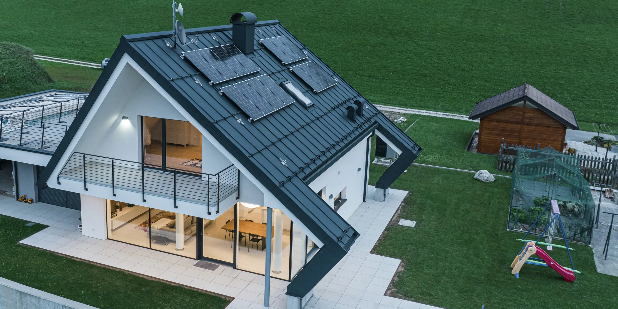Pohľad z vtáčej perspektívy na moderný rodinný dom v Borgo Valbelluna za súmraku. Svetlá osvetľujú obytné priestory v dome. Nápadná strecha s nezvyčajnými presahmi bola pokrytá robustnou strešnou krytinou PREFALZ vo farbe P.10 antracit. Odolná hliníková strecha a fotovoltické moduly spájajú udržateľnosť s kvalitným dizajnom. Nápadný presah strechy chráni fasádu a terasu pred poveternostnými vplyvmi. Veľké presklené plochy vytvárajú svetlom zaliatu obytnú atmosféru, zatiaľ čo obklopujúci balkón a priestranná terasa maximalizujú komfort bývania. Hliník PREFA je symbolom stability, elegancie a oslobodenia od údržby.
