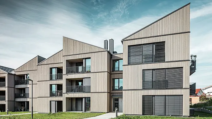 Vstupná časť budovy v areáli Schmucker v Utting am Ammersee. Architektúra zaujme jasnými líniami a nápadnými šikmými jednoplášťovými strechami, ktoré sú pokryté odolnými strešnými panelmi PREFA R.16 v tmavosivej farbe P.10. Elegantná drevená fasáda dokonale ladí s veľkými oknami a balkónmi, ktoré poskytujú dostatok svetla a priateľskú atmosféru. Trvalo udržateľný dizajn hliníkových výrobkov PREFA podčiarkuje inovatívnu hybridnú konštrukciu tohto architektonicky sofistikovaného rezidenčného projektu.
