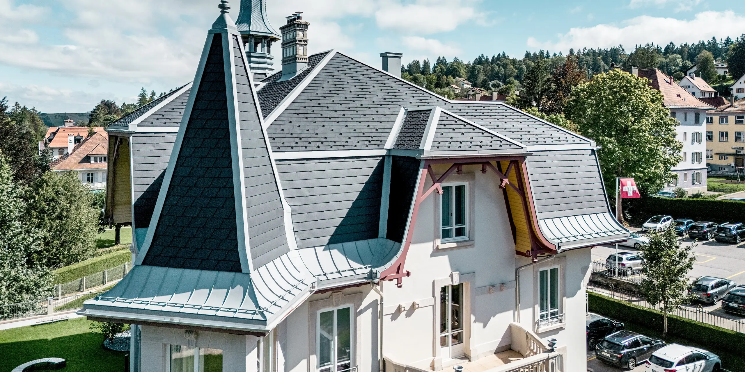 Faszinierende Detailansicht der Manufacture Horlogère du Locle in der Schweiz mit markantem Turm als architektonischem Highlight. Die vielschichtige Dachlandschaft beeindruckt durch steil geneigte Flächen und elegante Rundungen. PREFA Dachschindeln in P.10 Schwarz verleihen der Struktur klare Linien und ein dynamisches Relief, während PREFALZ in Patinagrau geschwungene Übergänge und komplexe Formen präzise umsetzt. Das leichte Aluminiumdach überzeugt durch Langlebigkeit, hohe Witterungsbeständigkeit und maximale Gestaltungsfreiheit bei anspruchsvoller Dacharchitektur.