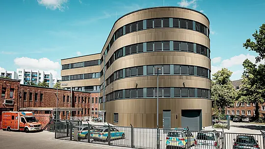 Výrazný exteriér policajnej stanice č. 43 v Hamburgu: Zakrivená nová budova s hliníkovou fasádou PREFA z eloxovaných extrudovaných profilov „Zacke“ v odtieni strednej bronzy pôsobí v mestskom prostredí ako výrazný prvok, ktorý priťahuje pozornosť. Jemná vertikálna štruktúra vytvára živé efekty svetla a tieňa a zdôrazňuje zaoblené tvary architektúry. V kontraste s ňou stojí historická tehlová budova. Hliník PREFA presviedča vysokou odolnosťou voči poveternostným vplyvom, ochranou proti korózii, dlhou životnosťou a minimálnymi nárokmi na údržbu – je ideálny pre udržateľné, funkčné a estetické riešenia fasád.