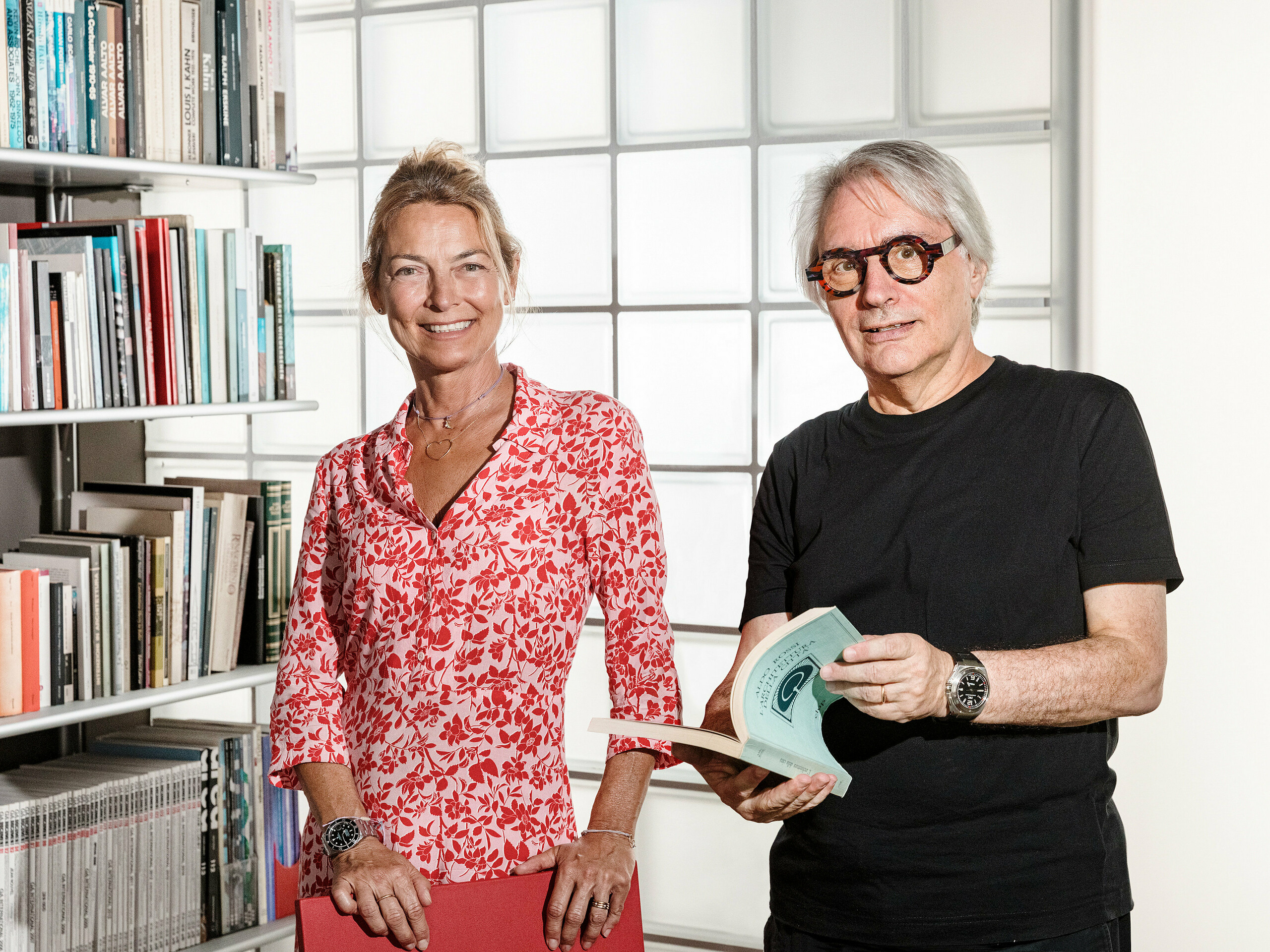 Architekt Claudio Lucchin z Bolzana a jeho kolegyňa Daniela Varnier pózujú pred modernou stenou zo sklenených tvárnic, ktorá zaujímavým spôsobom láme svetlo. Claudio Lucchin je celý oblečený v čiernom a drží v rukách knihu. Daniela Varnier má na sebe jasne červený top a priateľsky sa usmieva. Toto štýlové prostredie symbolizuje inováciu a transparentnosť. Scéna predstavuje futuristickú architektúru, ako bola realizovaná v kasárňach „Tempesti” v Corvare v Južnom Tirolsku.