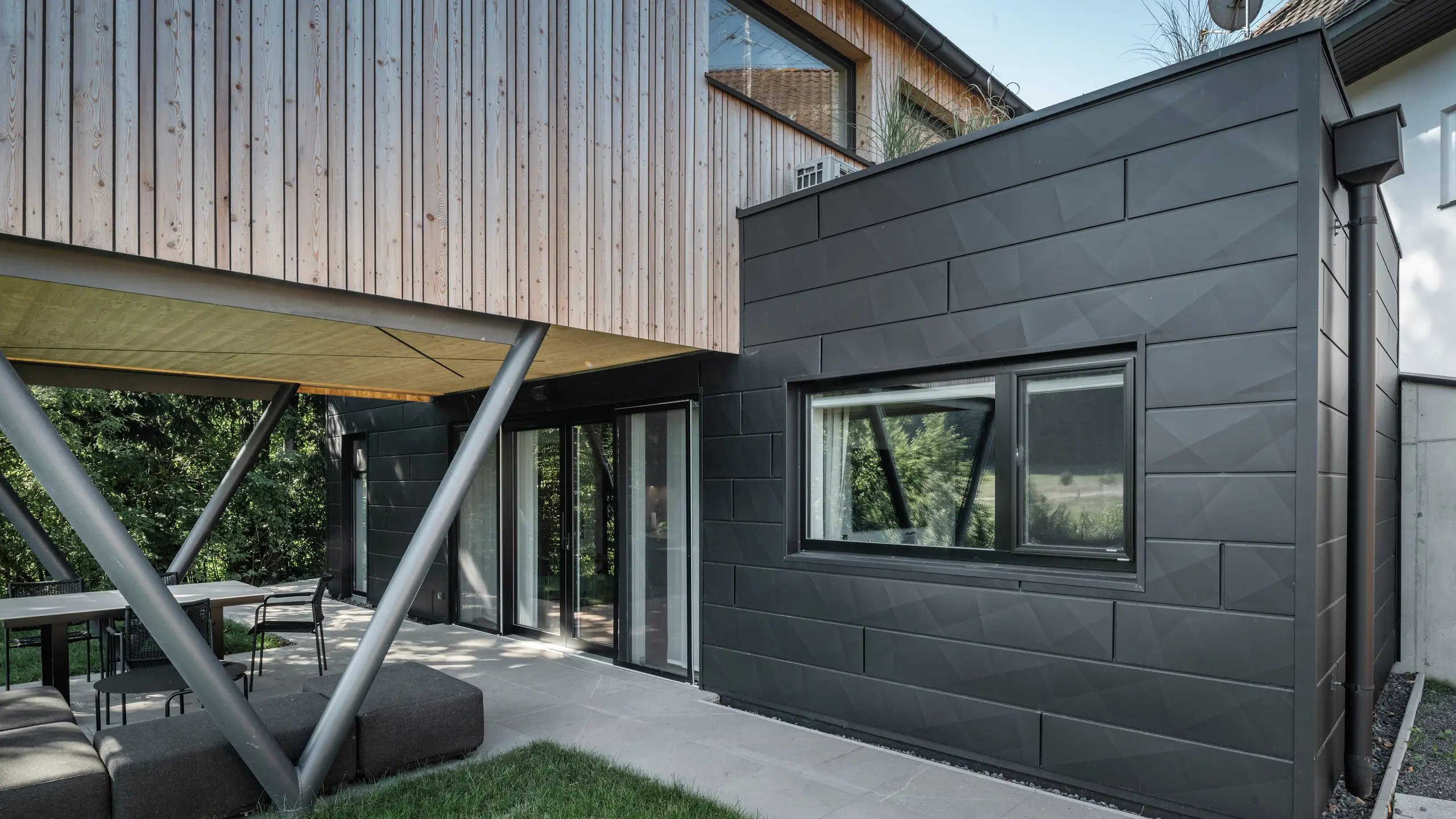 Architektonický highlight: „Woodstock Chalet 5“ v St. Martin am Innkreis, Horné Rakúsko. Moderné ubytovanie pre svadby je zdôraznené použitím antracitových fasádnych panelov PREFA Siding.X. Hliníkové prvky tvoria silný kontrast s jasnými drevenými panelmi a kovovými nosnými konštrukciami. Tmavé okenné rámy sa harmonicky začleňujú do dizajnu fasády, čím budova získava elegantný vzhľad. Táto fotografia ukazuje estetickú rovnováhu medzi prirodzenou textúrou dreva a industriálnym šarmom hliníkovej fasády, ktorá je typická pre dizajn moderných budov.