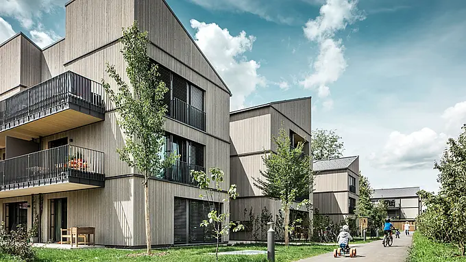 Moderné bytové domy na pozemku Schmucker v Utting am Ammersee, ktoré zaujmú inovatívnou hybridnou konštrukciou z dreva a udržateľnou architektúrou. Budovy sú vybavené odolnými strešnými panelmi PREFA R.16 a PREFALZ. Kombinácia elegantných drevených fasád a veľkých okien vytvára priateľskú a príjemnú atmosféru. Deti sa hrajú na zelených pozemkoch, ktoré prispievajú k harmonickému začleneniu do prírodného prostredia. Kvalitné materiály a premyslený dizajn podčiarkujú modernú a udržateľnú výstavbu tohto rezidenčného projektu.