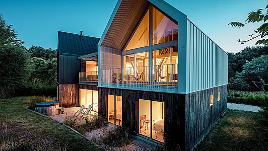 Moderný obytný súbor Le Rhune zaujme počas modrej hodinky výraznou architektúrou dvoch vzájomne posunutých objektov. Svetlý hliníkový plášť PREFALZ plynulo prepája strechu a fasádu predného domu do precízne tvarovaného celku a zvýrazňuje čistú geometriu sedlovej strechy. Veľkorysé presklené plochy vyžarujú do exteriéru teplé svetlo a vytvárajú pôsobivý kontrast s okolitou prírodou. Trvácny a poveternostným vplyvom odolný systém stojatej drážky PREFALZ umožňuje vytvárať filigránske detaily, súvislú obálku budovy a súčasnú interpretáciu tradičnej bretónskej architektúry.
