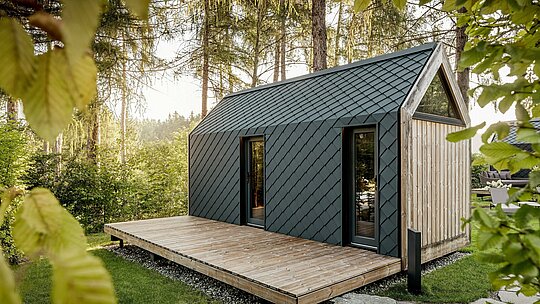 Minimalistický domček typu „tiny house“ v Secret Koi Garden pri jazere Natterer See sa nachádza uprostred pokojnej lesnej krajiny. Strecha a fasáda sú obložené malými hliníkovými strešnými a fasádnymi šablónami PREFA v odtieni P.10 antracitová, ktorých presná štruktúra pripomínajúca rybiu kožu jemne odráža slnečné lúče. Priestranná drevená terasa otvára priestor k prírode, zatiaľ čo odolný a poveternostne odolný hliník PREFA chráni architektúru a vytvára výrazný kontrast s drevom a zeleňou.