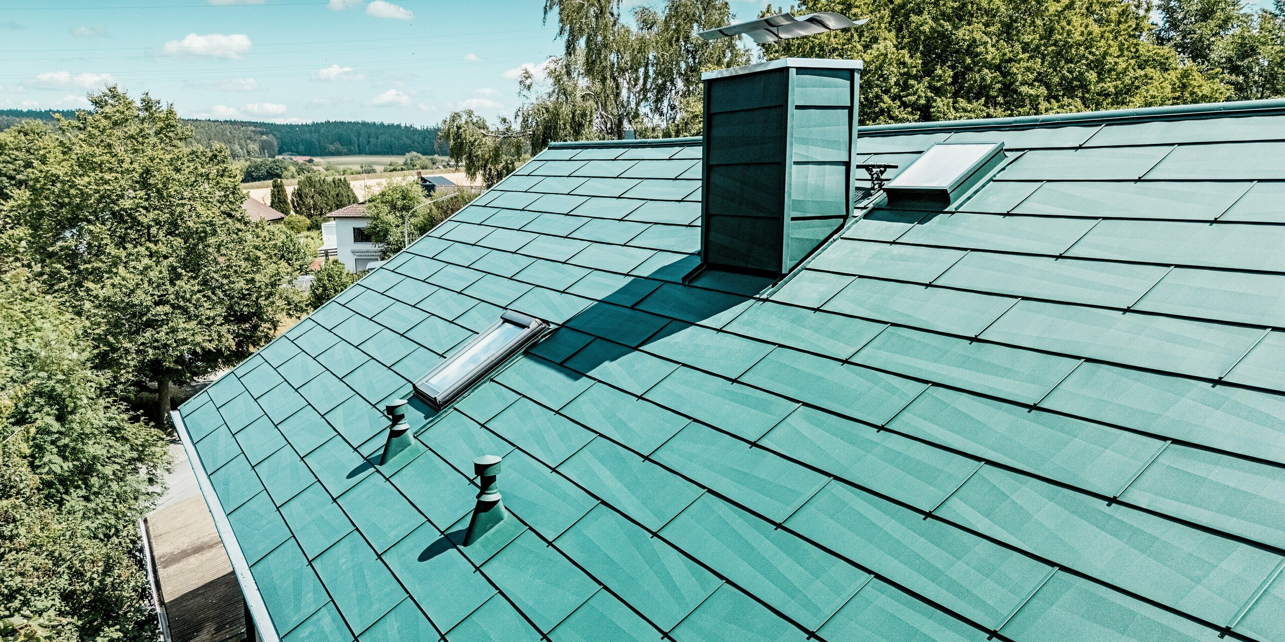 Modernisiertes Dach eines Einfamilienhauses in Schwarzenbach-Martinlamitz mit dem langlebigen PREFA Dachpaneel FX.12 in P.10 Moosgrün. Das detailreiche Bild zeigt das markante Aluminiumdach mit klaren Linien, strukturierter Oberfläche und integrierten Dachfenstern sowie einem formschön verkleideten Kamin. Die reflektierenden Lichtspiele auf den Paneelen unterstreichen das moderne Design. Dank der widerstandsfähigen Aluminiumoberfläche bietet das Dach optimalen Schutz vor Witterung, ist wartungsfrei und überzeugt durch eine stilvolle, zeitlose Ästhetik.