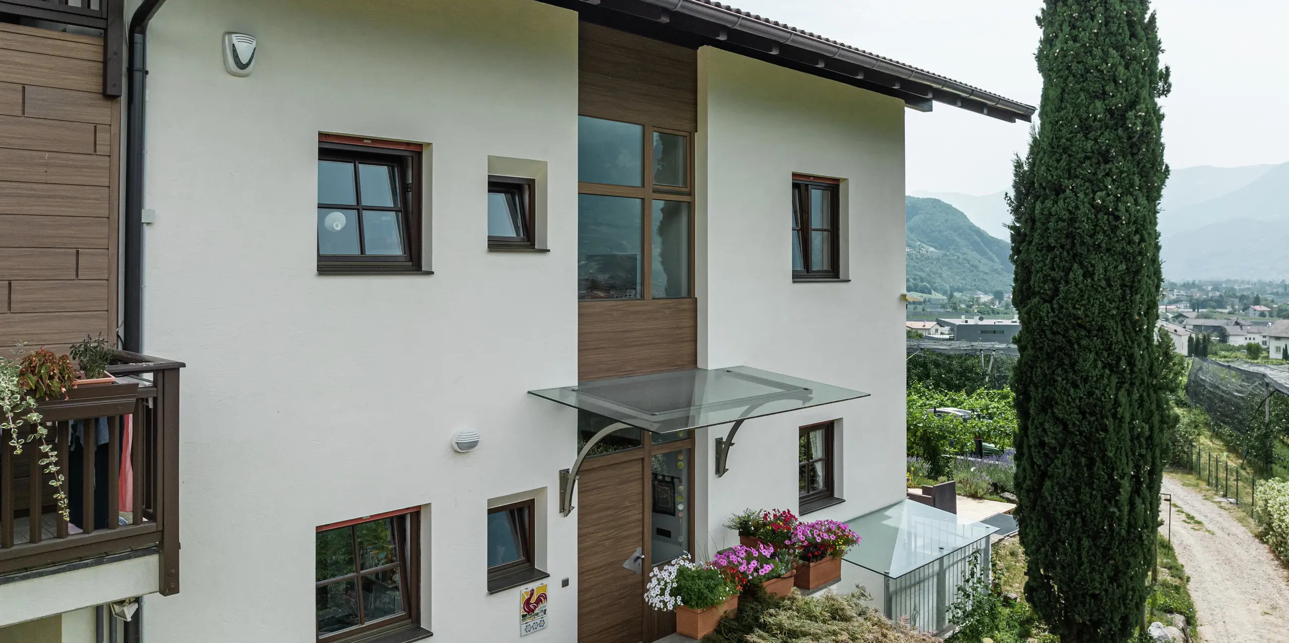 Vstupná časť rekreačných apartmánov Prechtlhof v Merane, Južné Tirolsko, s moderným dizajnom fasády z omietky a obkladov PREFA v orechovo hnedom vyhotovení. Bezúdržbová hliníková fasáda v drevenom vzhľade dopĺňa tradičný dizajn a zaisťuje trvácne a odolné riešenie voči poveternostným vplyvom.