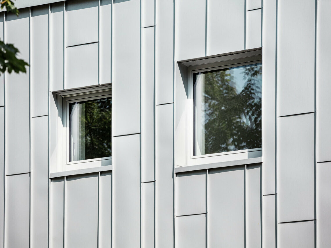 Detailný záber na modernú fasádu budovy s vertikálnymi striebornými kovovými hliníkovými panelmi Prefalz. Dve pravouhlé okná s tmavými rámami odrážajú zeleň stromov a rozbíjajú štruktúrovaný vzor fasády.