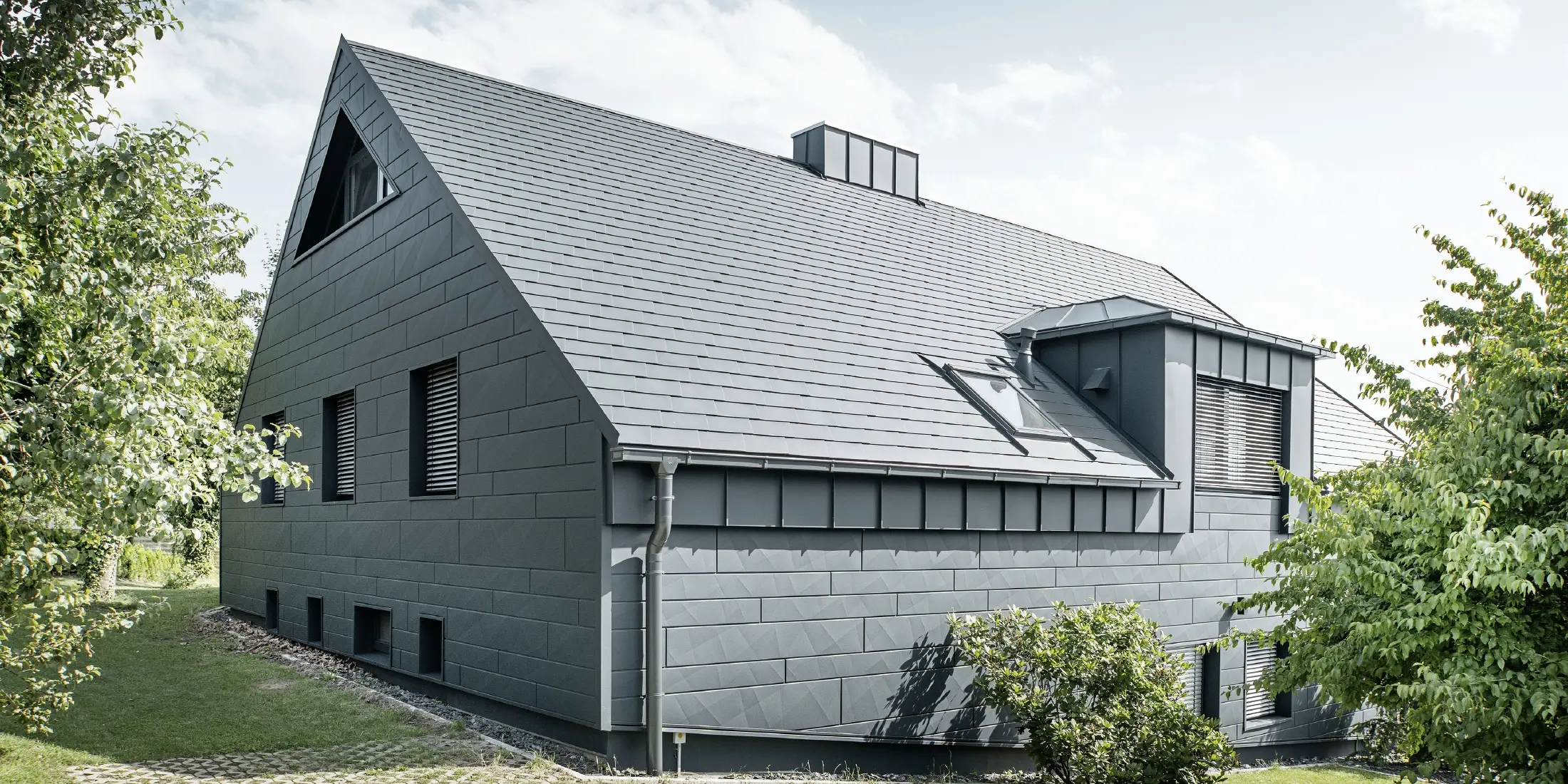 Veľký rodinný dom so sedlovou strechou a mnohými oknami. Celý dom bol obložený hliníkovým plášťom v antracitovej farbe, na streche boli použité hliníkové strešné šindle PREFA a na fasáde Siding.X v antracitovej farbe.