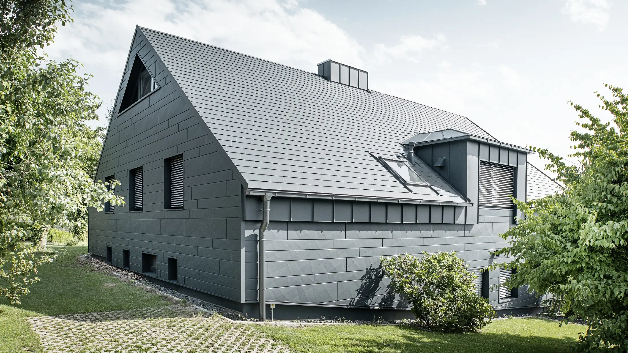 Veľký rodinný dom so sedlovou strechou a mnohými oknami. Celý dom bol obložený hliníkovým plášťom v antracitovej farbe, na streche boli použité hliníkové strešné šindle PREFA a na fasáde Siding.X v antracitovej farbe.