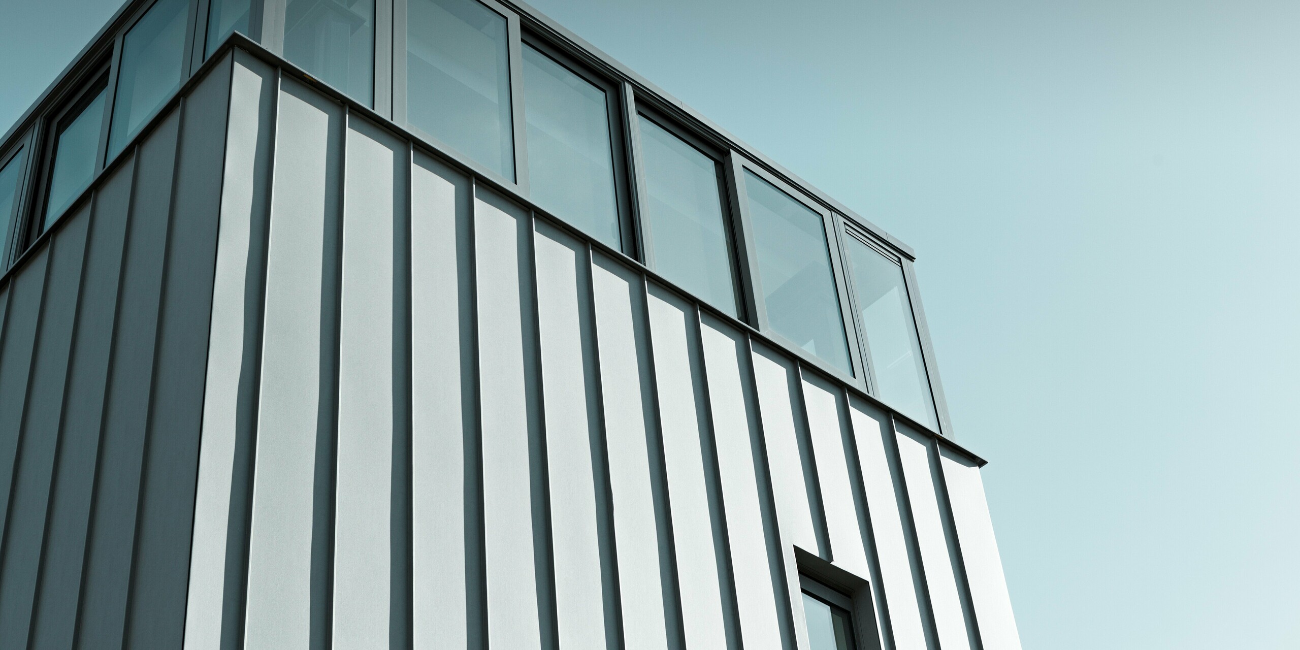 Dynamický detailný záber modernej fasády PREFALZ v patinovo šedej farbe. Čisté línie vertikálnych stojatých falcov sa stretávajú s veľkorysým zasklením v hornom poschodí, ktoré vytvára svetlo a transparentnosť. Odolná hliníková fasáda PREFA zaisťuje trvalú ochranu a architektonicky vytvára výrazný dojem.