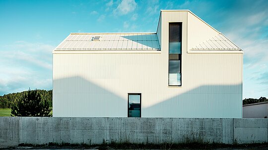 Moderný rodinný dom vo Freistadte bol fotografovaný z bočného pohľadu, aby vynikol výrazný dizajn fasády a strechy. Čisto biele hliníkové obklady PREFALZ v kombinácii so strmou sedlovou strechou a úzkymi oknami dodávajú budove moderný a nadčasový vzhľad. Vertikálne drážky fasády dodávajú budove dynamický, textúrovaný vzhľad. Hra svetla, ktoré dopadá na fasádu, vytvára zaujímavé tieňové efekty a zdôrazňuje presné línie dizajnu. Táto čistá a minimalistická architektúra je podčiarknutá odolnou a poveternostne odolnou fasádou PREFALZ, ktorá je známa svojou trvanlivosťou a ľahkou údržbou.
