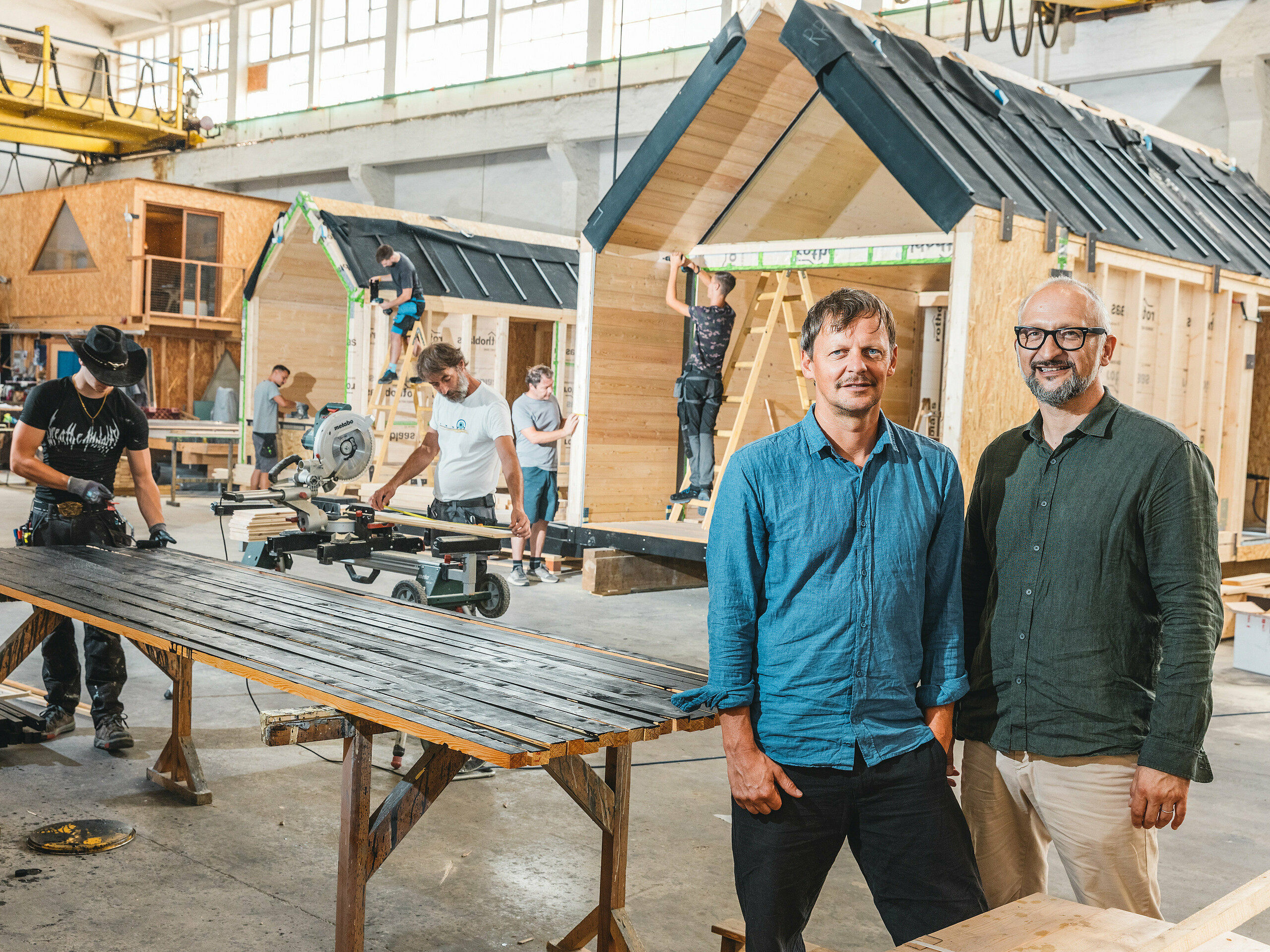 Vo výrobnej hale vznikajú minimalistické minidomy spoločnosti Lushna. Táto firma je úspešná na medzinárodnej úrovni a špecializuje sa na minidomy v prírodných rezortoch. V popredí sú obaja zakladatelia firmy: architekt Peter Ličen a marketingový špecialista Jaka Ažman. V pozadí sa montujú domy typu A-Frame. Výrazný kontrast dreva a tmavého hliníka robí z tejto architektúry vizuálny magnet, o čom svedčí Secret Koi Garden pri jazere Natterer See.