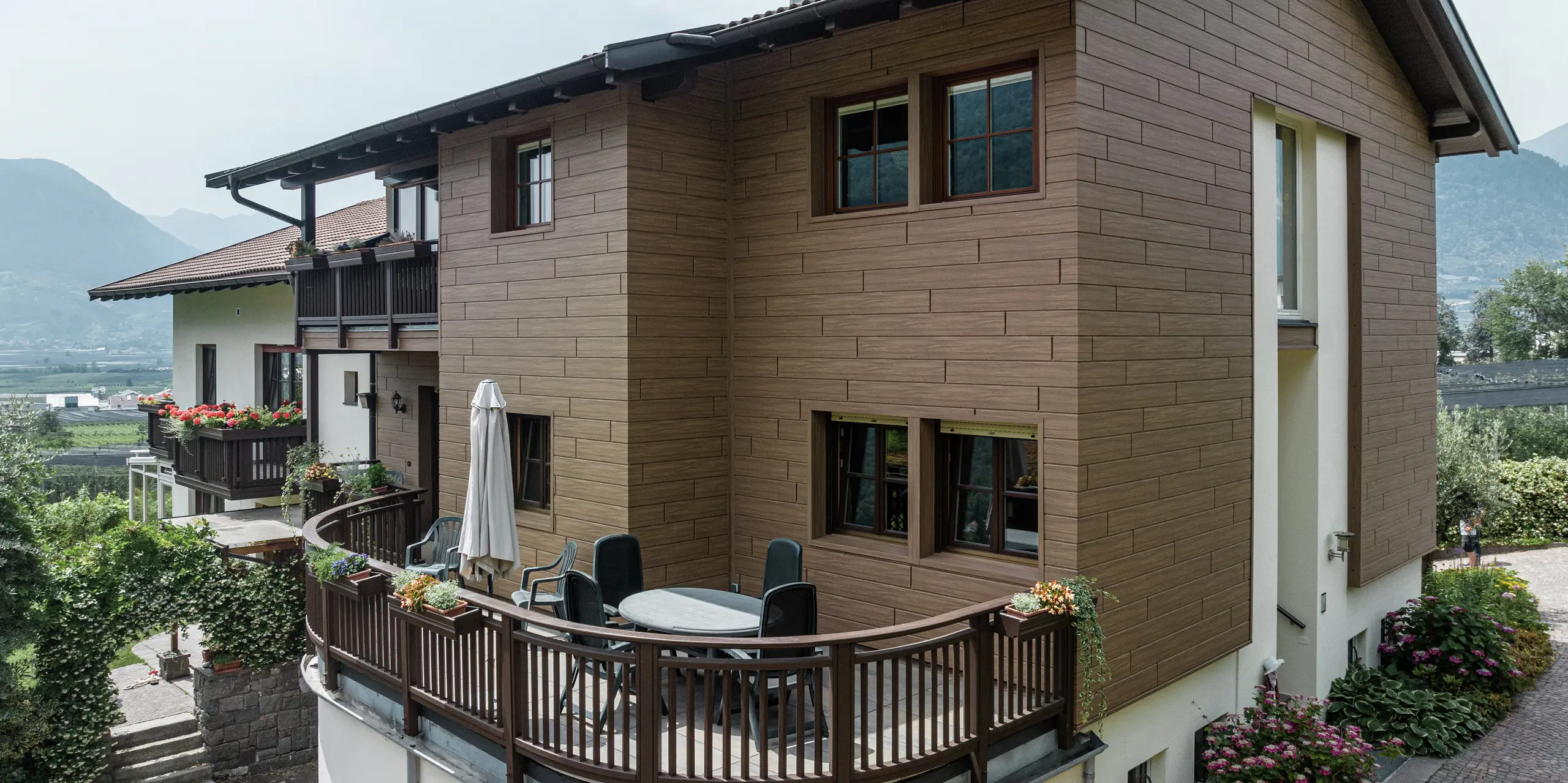Renovované apartmány Prechtlhof v Merane, Južné Tirolsko, s hliníkovými obkladmi PREFA Siding v odtieni vlašský orech hnedý. Bezúdržbová hliníková fasáda v drevenom vzhľade spája modernú estetiku s tradičným šarmom a harmonicky zapadá do alpského prostredia. Okrúhly balkón v popredí láka k oddychu v nádhernej južnotirolskej krajine.