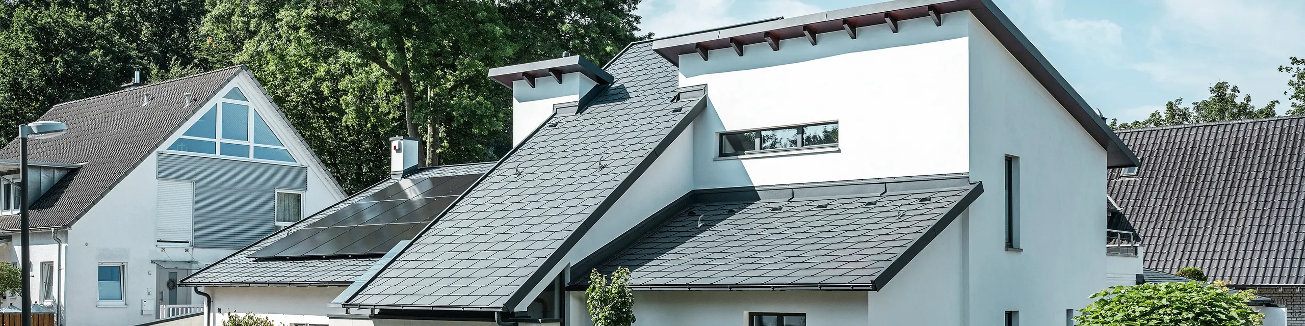 Moderný rodinný dom s bielou omietkovou fasádou a tmavou šikmou strechou PREFA z hliníka. Na streche sú integrované fotovoltaické moduly a hliníkové odvodňovacie systémy v antracitovej farbe. Budova sa nachádza v udržiavanej obytnej štvrti so zelenými predzáhradkami, dláždenou príjazdovou cestou a okolitými rodinnými domami.