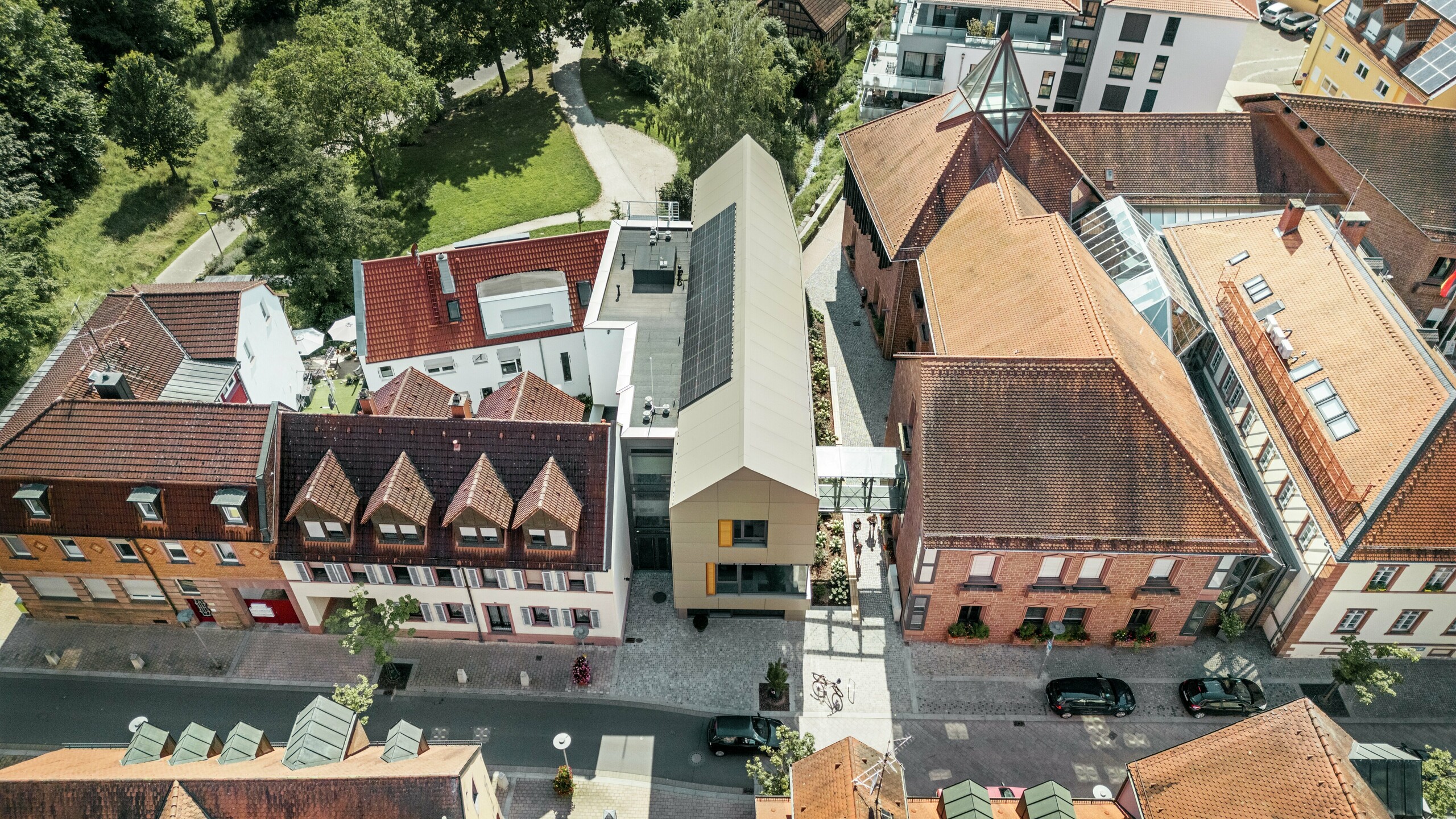 Radnica v nemeckom Alzenau sa nachádza v centre historického mestského prostredia, obklopená tradičnými budovami s červenými škridlovými strechami. V kontraste s nimi stojí moderná radnica, ktorej fasáda je obložená hliníkovými kompozitnými panelmi PREFA v elegantnom bronzovom odtieni. Inovatívna architektúra sa jasne odlišuje od susedných klasických budov, ale napriek tomu sa harmonicky začleňuje do mestského prostredia.