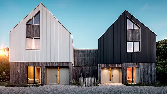 Pohľad z prednej strany na obytný súbor Le Rhune odhaľuje dva vzájomne posunuté objekty s identickým architektonickým výrazom, ktoré sa od seba odlišujú kontrastným farebným riešením v bielej a čiernej. Čisté tvary sedlových striech a súvislý hliníkový plášť PREFALZ dodávajú budovám sochársky charakter a pôsobivo zvýrazňujú precízne línie stojatej drážky. Teplé svetelné akcenty prenikajúce cez veľkorysé presklené plochy vytvárajú výrazný kontrast k minimalistickej architektúre. Odolný a trvácny hliník PREFALZ spája strechu a fasádu do modernej, harmonickej obálky budovy s vysokou estetickou a architektonickou hodnotou.