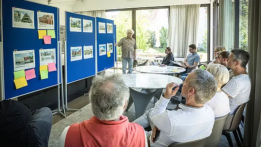 PREFARENZEN 2027 Dialóg odborníkov v svetlej, modernej seminárnej miestnosti: Moderátor Matthias Finkentey stojí pred modrými nástenkami, na ktorých sú prezentované architektonické projekty s hliníkovými riešeniami PREFA. Porota pozorne sedí pri okrúhlych stoloch, diskutuje a zapisuje si dojmy. Veľké okná vpúšťajú dovnútra denné svetlo, zatiaľ čo vonku je vidieť zelenú záhradu. Scéna znázorňuje prepojenie kreatívnej diskusie o architektúre, funkčného plánovania a hodnotenia dizajnu. Hliníkové produkty PREFA sú synonymom pre trvácne, estetické strechy a fasády, ktoré stelesňujú inováciu, kvalitu a inšpiráciu v architektonických projektoch.