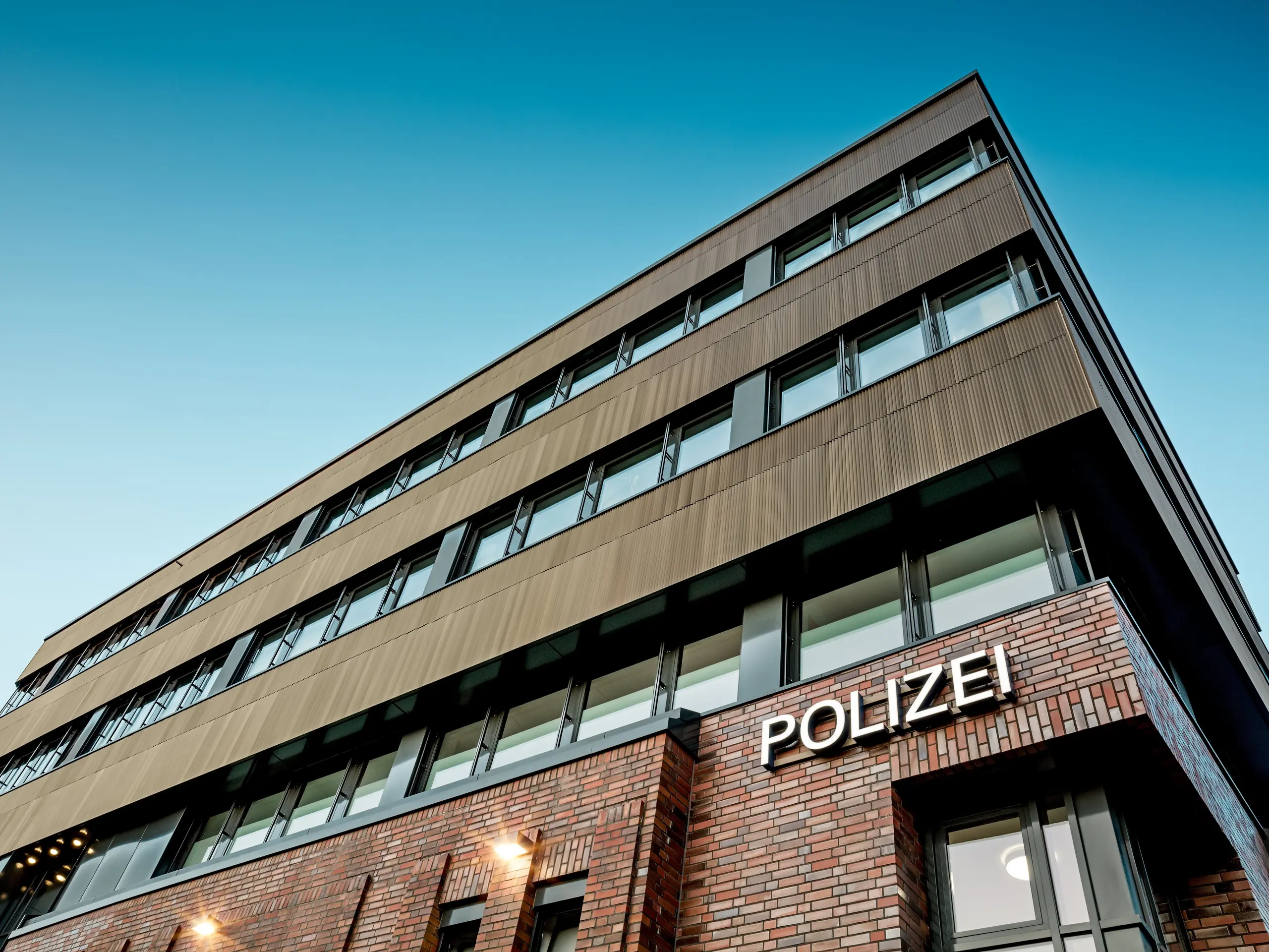 Architektonický pohľad zospodu na policajnú stanicu č. 43 v Hamburgu: Nad pôvodnou tehlovou budovou sa týči precízne členená nová stavba s hliníkovou fasádou PREFA z extrudovaných profilov „Zacke“ v elegantnom odtieni strednej bronzy. Jemne rýhovaný povrch púta pozornosť a vytvára fascinujúce odlesky svetla pozdĺž čistých, horizontálnych pásov okien. Hliník PREFA je synonymom najvyššej odolnosti proti korózii, dlhej životnosti a minimálnej údržby – ide o udržateľné riešenie s výrazným dizajnom pre modernú a funkčnú fasádnu architektúru.